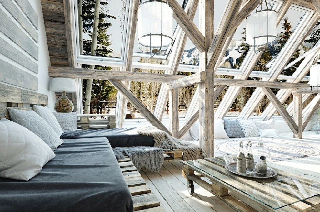 Sunlit A-frame mountain living room with reclaimed-timber cross beams, pallet-wood daybeds and coffee table, and a wall of windows opening onto snowy pines and peaks.